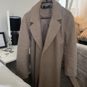 Beige kappa från Vero Moda - Säljer en stilren beige kappa från Gina i mycket bra skick. Perfekt för höst och vår med sin klassiska design och långa ärmar. Kappan har en elegant krage och är gjord i ett mjukt material som känns lyxigt. Passar både till vardags och fest!