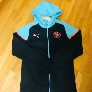 Manchester City overall - Säljer en snygg svart och blå hoodie från Puma med dragkedja och huva. Den har ett coolt tryck på bröstet och är perfekt för både träning och vardag. Den är i mycket bra skick och redo för nya äventyr! 💙