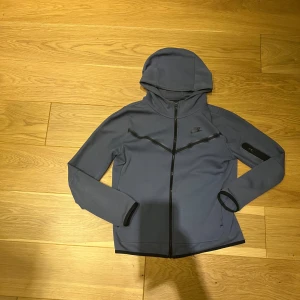 Nike tech fleece  - Säljer en blå Nike tech fleece kofta från Nike i mycket bra skick. Den har en dragkedja framtill och en snygg, sportig design med långa ärmar. Perfekt för träning eller vardagsbruk. Huvan ger extra skydd mot väder och vind. Passar perfekt för höst och vår!