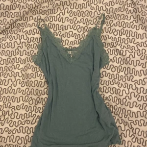 Grön ribbad linne med spetsdetaljer - Säljer en superfin grön ribbad linne med spetsdetaljer. Den har smala axelband och en v-ringad design med söta rosetter framtill. Perfekt för sommardagar eller som ett lager under en kofta. Passar både till vardags och festliga tillfällen! 