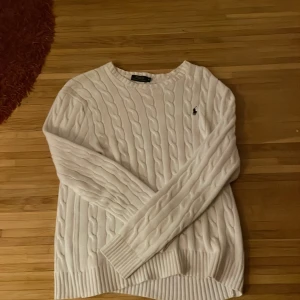 Vit stickad tröja från Ralph Lauren - Säljer en klassisk vit stickad tröja från Ralph Lauren med kabelstickat mönster. Den har en broderad logga på bröstet och är i mycket bra skick. Perfekt för både höst och vinter, och den är supermysig att ha på sig. Passar till alla tillfällen!