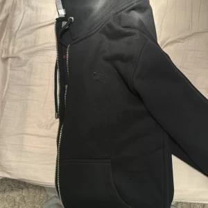 Svart huvtröja från Burberry - Säljer en snygg svart huvtröja från Burberry i mycket bra skick. Den har en dragkedja framtill och en klassisk rutig insida i huvan. Perfekt för höst och vinter, med långa ärmar och en bekväm passform. Passar både till vardags och lite finare tillfällen. Helt som ny, fleece på hela tröjan innanför. Hör av er vid frågor!