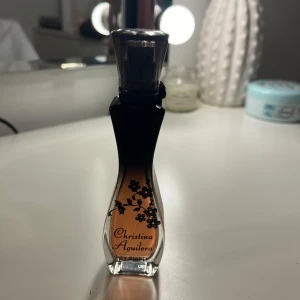 Parfym från Christina Aguilera - Säljer en elegant parfymflaska från Christina Aguilera. Flaskan har en smal, kurvig form med en svart kork och ett vackert blommönster. Färgen går från svart till en varm bärnsten. Perfekt för en kväll ute eller som en fin present!
