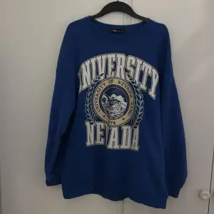 Säljer en blå sweatshirt med stort University of Nevada-tryck på framsidan. Tröjan är i bra skick och perfekt för en avslappnad stil. Den har långa ärmar och är gjord i ett mjukt material som känns skönt mot huden. Perfekt för höst och vinter!