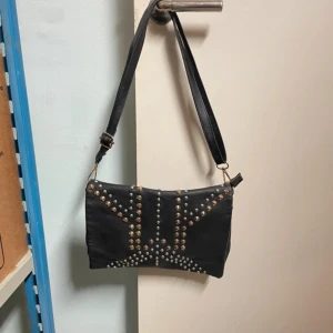 Svart axelväska med nitar - Snygg svart axelväska i skinn med nitar på framsidan. Väskan har en dragkedja upptill och en justerbar axelrem. Passar både till vardags och fest.