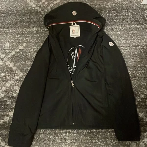 Svart jacka från Moncler - Säljer en snygg svart jacka från Moncler i mycket bra skick. Den har en dragkedja framtill och en huva med Moncler-logga. Perfekt för höst och vår, med en stilren design och hög kvalitet. Passar både till vardags och lite finare tillfällen.