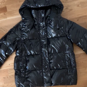 Glossy Jacket Calvin Klein Jeans - Svart pufferjacka från Calvin Klein Jeans i storlek M. Jackan är glansig och har en huva samt dragkedja framtill.                                                                                   •Skick: Använd endast ett par gånger ser helt ny ut.                                                               •Storlek:  M  passar för storlek S också