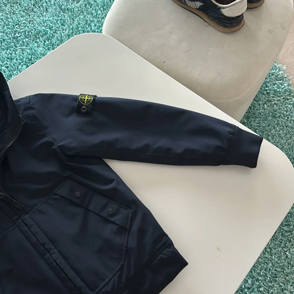 Säljer en snygg blå jacka från Stone Island Junior. Den har en dragkedja framtill och långa ärmar. Perfekt för höst och vår. Jackan är i mycket bra skick och har en cool logga på ärmen. Passar perfekt för dig som vill ha en stilren look!. Jackor.