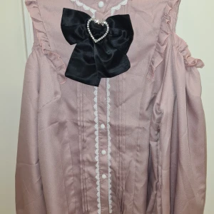 Jirai Kei blus med svart rosett - Säljer en superfin rosa jirai kei blus med en stor svart rosett framtill. Perfekt för höst och vår med sina romantiska detaljer och volanger. Den har också en söt hjärtformad detalj i mitten av rosetten.