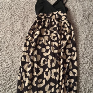Svart och beige leopardmönstrad klänning - Snygg klänning med svart överdel och leopardmönstrad kjol i beige och svart. Tunna axelband med en smickrande passform. Perfekt för en festlig kväll eller en utekväll med vänner. Klänningen har en omlott-look och är gjord i ett mjukt och bekvämt material. Klänningen passar både M och S