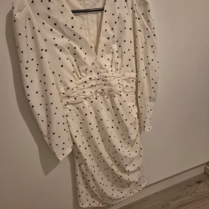Vit klänning med hjärtmönster - Säljer en superfin vit klänning med små svarta hjärtan. Den har en v-ringad design och långa ärmar. Klänningen är perfekt för både vår och sommar, och den har en smickrande passform med rynkdetaljer i midjan. Perfekt för en romantisk kväll eller en festlig tillställning! 💖