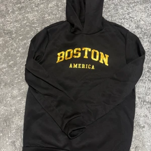 Svart hoodie med Boston-tryck - Vi Säljer en svart hoodie med guldfärgat tryck 'BOSTON AMERICA' på bröstet. Perfekt för en avslappnad stil och håller dig varm under kyliga dagar. Den har en klassisk passform med långa ärmar och en bekväm huva. Passar både till vardags och chillkvällar.