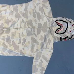 Vit och grå camo hoodie från A Bathing Ape - Säljer en snygg vit och grå camo hoodie från A Bathing Ape i storlek S. Den har WGM-broderi på sidan. En liten tråd som sticker ut vid ögat men lätt att fixa. Annars så är den bra kvalitet. Skriv vid frågor och pris går att diskutera vid snabb affär🔥🔥