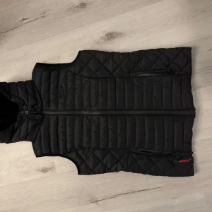 Svart dunväst från Marikoo - Säljer en snygg svart dunväst från Marikoo i storlek M. Västen har en quiltad design med dragkedja framtill och två praktiska fickor. Perfekt för höst och vinter, håller dig varm och stilren. Passar både till vardags och lite mer uppklädda tillfällen. 🖤