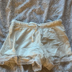 Beige shorts med spetsdetaljer - Säljer ett par söta beige shorts med spetsdetaljer i nederkant. De har en bekväm resårmidja med knytband för justerbar passform. Perfekta för en avslappnad sommardag eller mysiga stunder hemma. 🩳✨