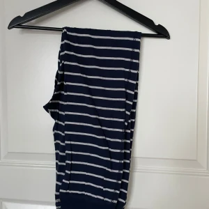 Randiga pyjamasbyxor med spetsdetaljer - Säljer ett par snygga randiga pyjamasbyxor i mörkblått och vitt. De har en bekväm passform med elastisk midja och snörning. Byxorna är dekorerade med fina spetsdetaljer vid fickorna och midjan. Perfekta för en avslappnad hemmakväll!