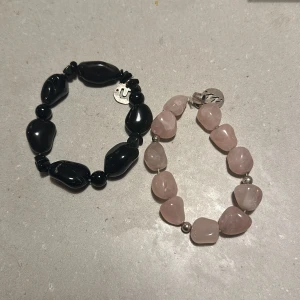 Två armband med stenar - Säljer två snygga armband med stenar. Det ena är svart med glansiga, oregelbundna stenar och små silverdetaljer. Det andra är rosa med mjuka, rundade stenar och silverpärlor emellan. Perfekta för att ge en bohemisk touch till din outfit!