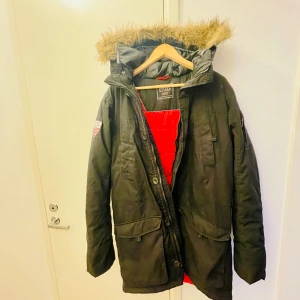 Svart parkajacka med pälsdetaljer - En svart parkajacka i mycket bra skick. Jackan har en mysig pälsfodrad huva och röd insida som ger en snygg kontrast. Jackan har sex fickor inuti /utåt. Den har långa ärmar och praktiska fickor framtill. Perfekt för kalla vinterdagar! ❄️