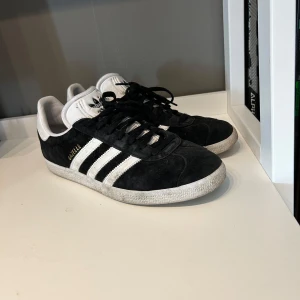 Adidas Gazelle i svart och vitt - Säljer ett par klassiska Adidas Gazelle i svart mocka med vita detaljer och snörning. De har den ikoniska tre-randiga designen och Adidas-loggan på hälen. Perfekta för  vardag. Pris kan diskuteras vid en snabb affär 
