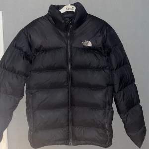 Säljer en svart dunjacka från The North Face i mycket bra skick. Jackan är perfekt för kalla vinterdagar och har en dragkedja framtill. Den är långärmad och har en klassisk pufferstil som håller dig varm och bekväm. Passar både till vardags och utomhusaktiviteter.