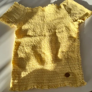 Gul ribbad topp med korta ärmar - Säljer en söt gul ribbad topp i storlek 134/140. Materialet är stretchigt, så passar för lite större storkekar. Har korta ärmar och ett gulligt märke med ett leende ansikte. Perfekt för sommardagar eller som en färgglad detalj i garderoben. Toppen är i bra skick och redo för nya äventyr! 🌞