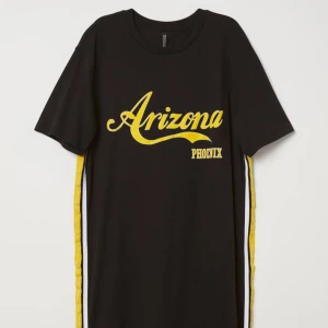 Slutsåld Svart klänning med Arizona-tryck - Snygg svart klänning med gult Arizona-tryck och texten Phoenix. Den har korta ärmar och gula samt vita ränder längs sidorna. Perfekt för en avslappnad dag eller en sportig look. Klänningen är i ett mjukt material som känns skönt mot huden. 100% bomull.  Skriv för fler bilder eller frågor 🥰