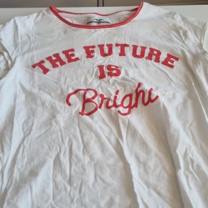 Vit t-shirt från Hampton Republic - Säljer en vit t-shirt från Hampton Republic med texten 'The Future is Bright' i rött. T-shirten har korta ärmar och röd kant vid halsen. Den är i bomull och har några små hål, men är fortfarande i acceptabelt skick. Perfekt för en avslappnad stil! Med tyvärr två små hål.