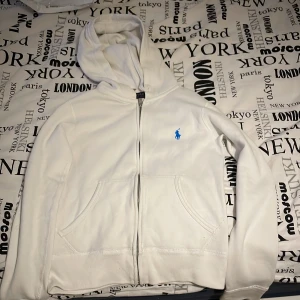 Vit hoodie från Ralph Lauren - Säljer en snygg vit hoodie från Ralph Lauren i bra skick. Den har en dragkedja framtill och en klassisk blå logga på bröstet. Perfekt för både vår och höst, med en skön passform och praktiska fickor den är även tjockare perfekt nu till vintertider inga defekter och endast använd vid 3 tillfällen . Hör av dig vid intresse!