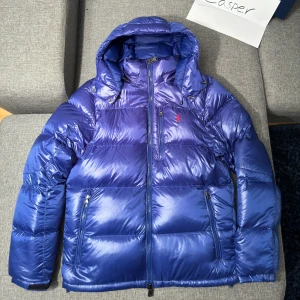 Blå dunjacka från Ralph Lauren - Säljer en snygg blå dunjacka från Ralph Lauren. Den har en avtagbar huva och är perfekt för kalla dagar. Jackan har en dragkedja framtill och två praktiska fickor. Den röda loggan på bröstet ger en extra touch av stil. Nyskick och aldrig använd, med prislappen fortfarande kvar. Perfekt för vintern! 💙