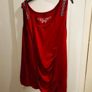 Röd ärmlös topp från Ze-Ze - Snygg röd ärmlös topp från Ze-Ze i storlek XL. Den har coola nitar på axelbanden och ett draperat tyg framtill som ger en unik look. Perfekt för en utekväll eller fest! 💃