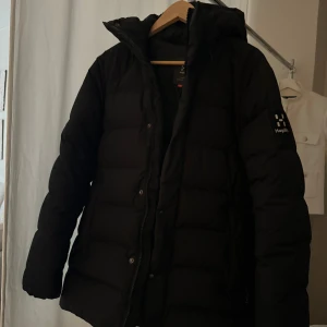 Svart dunjacka från Haglöfs - Säljer en svart dunjacka från Haglöfs i storlek S. Jackan är perfekt för kalla vinterdagar med sin varma och vadderade design. Den har långa ärmar och en praktisk dragkedja framtill. Märket Haglöfs syns tydligt på ärmen. Perfekt för dig som vill hålla dig varm och stilren i vinter! ❄️