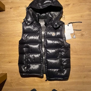 Svart dunväst från Moncler - Säljer en snygg svart dunväst från Moncler i mycket bra skick. Västen har en quiltad design med dragkedja och en avtagbar huva. Perfekt för höst och vinter, håller dig varm och stilren. Storlek 5, vilket motsvarar herr XL. Passar både till vardags och festliga tillfällen.