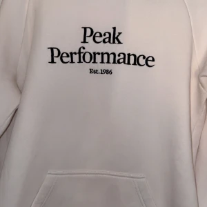 Vit hoodie från Peak Performance - Säljer en stilren vit hoodie från Peak Performance med svart text på bröstet. Hoodien har en klassisk design med känguruficka och justerbar huva med dragsko. Perfekt för en avslappnad look.