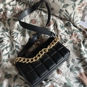 Svart quiltad axelväska med guldkedja - Snygg svart quiltad axelväska med en iögonfallande guldkedja. Perfekt för att ge din outfit en elegant touch. Väskan har en justerbar axelrem för bekväm passform.