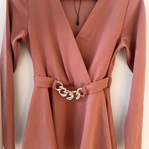 Rosa blus med kedjedetalj - Elegant rosa blus med v-ringning och långärmade. Blusen har en dekorativ kedjedetalj i midjan som ger en extra touch. Perfekt för en stilren look.