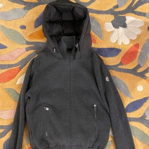 Stickad moncler cardigan - Snygg stickad svart/grå jacka från Moncler med quiltad design och dragkedja. Jackan har en hög krage, perfekt för kyliga dagar. Den har långa ärmar och ribbade muddar för extra komfort. Den är XL men moncler har mindre än vanliga storlekar och sitter som M/L. Den har inga deffekter. Priset är inte hugget i sten, fråga vid minsta fundering!😁