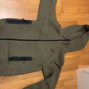 Olivgrön hoodie från Nike - Säljer en olivgrön hoodie från Nike med dragkedja och huva. Jackan har svarta detaljer och praktiska fickor med dragkedjor. Perfekt för en sportig look eller avslappnade dagar.