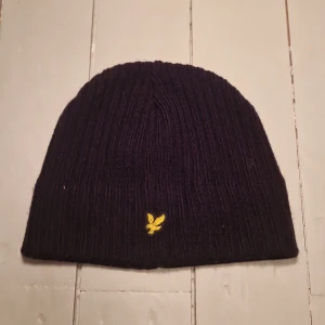 Svart ribbad mössa från Lyle & Scott - Snygg svart ribbad mössa från Lyle & Scott med ett gult broderat emblem framtill. Perfekt för att hålla värmen under kyliga dagar. Passar alla tack vare one size-designen.