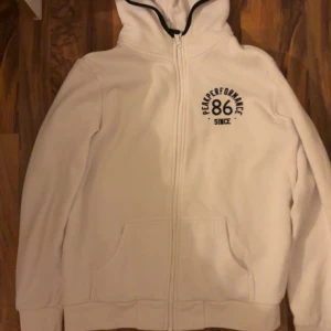 Vit hoodie från Peak Performance - Säljer en vit hoodie från Peak Performance med dragkedja och tryck på bröstet. Den har en bekväm passform och en praktisk huva med svart kant. Knappt använd och inga defekter, skriv gärna för frågor😊