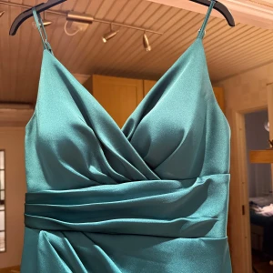 Grön långklänning från Kenza - Elegant turkos långklänning från Kenza i satin med omlott-design och smala axelband. Klänningen har en v-ringad urringning och en stilren slits framtill. Perfekt för speciella tillfällen ex bal, bröllop osv. Använd några gånger på bröllop