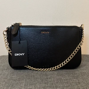 Svart axelväska från DKNY - Helt ny snygg svart axelväska från DKNY i skinn med guldfärgad kedja och dragkedja. Väskan har en avtagbar axelrem och en elegant design som passar till många tillfällen. Perfekt för en stilren look. Mått: 21x13x5cm. Kan sänka pris vid snabbköp!