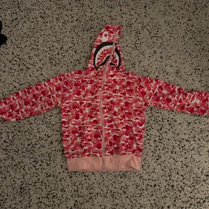 Rosa camo hoodie med hajmotiv - Cool hoodie i rosa camouflagemönster med hajmotiv på huvan. Den har en dragkedja framtill och långa ärmar. Perfekt för en unik och trendig stil.