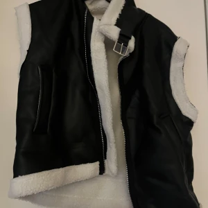 Svart skinnväst med vit pälsfoder - Snygg svart skinnväst med vit pälsfoder och dragkedja framtill. Västen har en cool spänndetalj vid halsen och är perfekt för en trendig look.