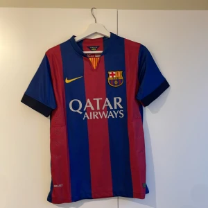 Blå och röd FC Barcelona fotbollströja från Nike - Säljer en officiell FC Barcelona fotbollströja från Nike i blått och rött med klubbens emblem och Qatar Airways-logga. Tröjan är kortärmad och har Dri-FIT-teknologi för optimal komfort. Perfekt för alla Barça-fans! 2014/15 hemma tröja 💙❤️