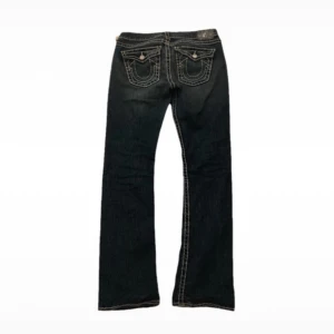 Lågmidjade True religion jeans - Säljer ett par vintage låg midjade true religion jeans. De är raka/ bootcut med billy fat stitch modell. Sitter som S/M och är i bra skick förutom det på låret. Vet inte om det är en del av designen eller inte. Midjemått: 42 cm Innerbenslängden: 84 cm