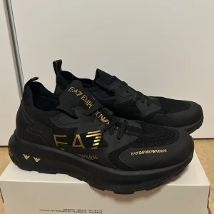 Svarta sneakers från EA7 Emporio Armani.  - Snygga svarta sneakers från EA7 Emporio Armani med guldiga detaljer och logotyp. Skorna har en modern design med storlek 42 2/3