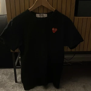 Svart Cdg t-shirt - Säljer en svart t-shirt från Comme des Garçons med det ikoniska röda hjärtat på bröstet. T-shirten är kortärmad och har en klassisk passform. Perfekt för en stilren look.