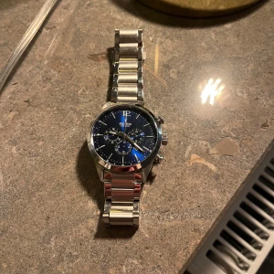 Silverfärgad klocka med blå urtavla från Festina - Snygg klocka från Festina med en stilren blå urtavla och silverfärgad metallarmband. Klockan har tre mindre urtavlor och en kronograf-funktion. Perfekt för den som gillar en klassisk och elegant stil.