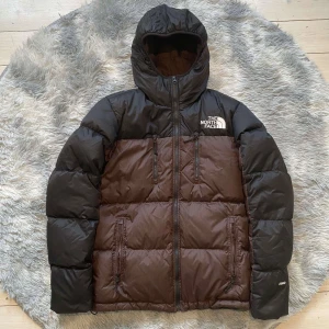 Brun och svart dunjacka från The North Face - Säljer en snygg brun och svart dunjacka från The North Face med huva och dragkedja. Jackan har broderad logga på bröstet och är perfekt för kalla dagar. Den är långärmad och har en klassisk pufferdesign.