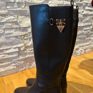 Svarta stövlar från Guess - Snygga svarta stövlar från Guess i skinn med dekorativt metallspänne och spetsig tå. Perfekta för en stilren look.